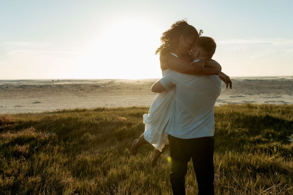 A couple embraces on a hilltop at sunset, silhouetted against the golden sky and grassy meadow.