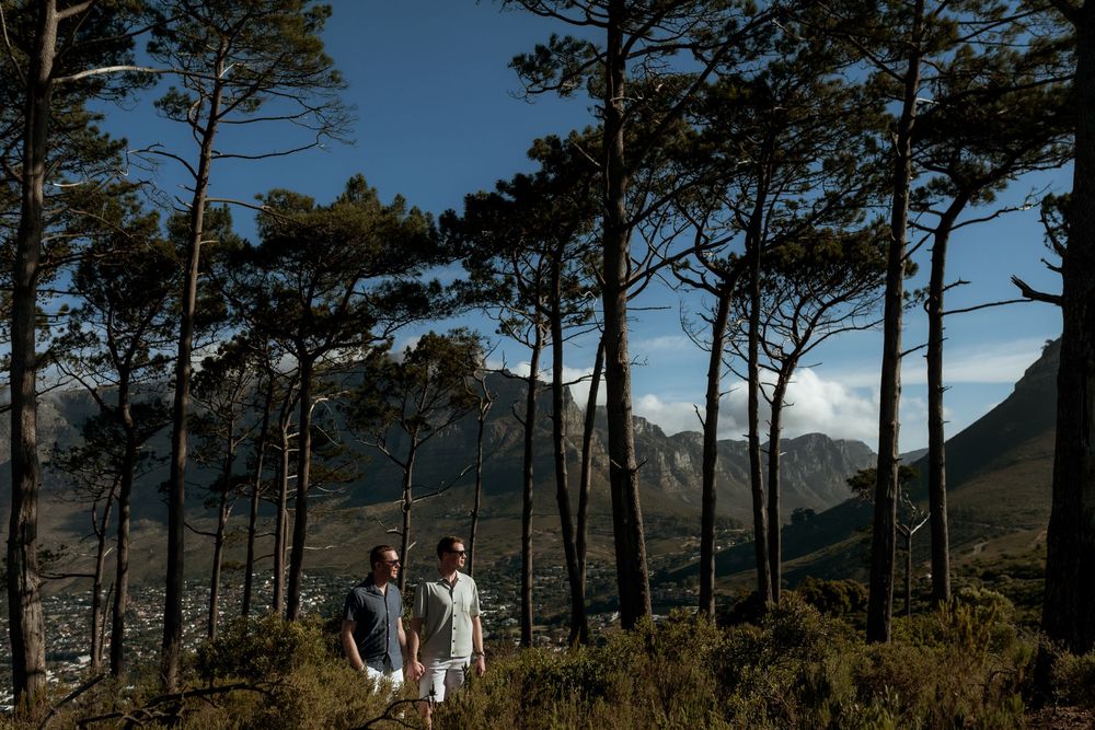 Two figures stand among tall pine trees on a mountainside with dramatic mountain peaks in the distance.