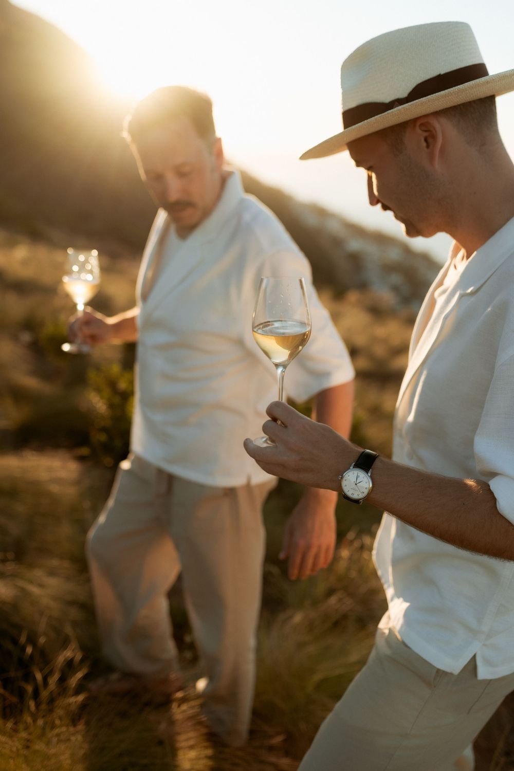 A couple in white clothing stands together during golden hour sunset on a hillside.