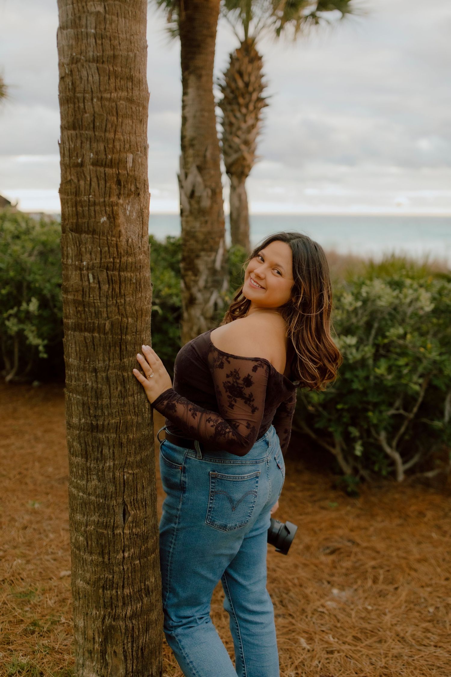 Woman in dark lace off-shoulder top and jeans hugging palm tree near ocean on cloudy day.