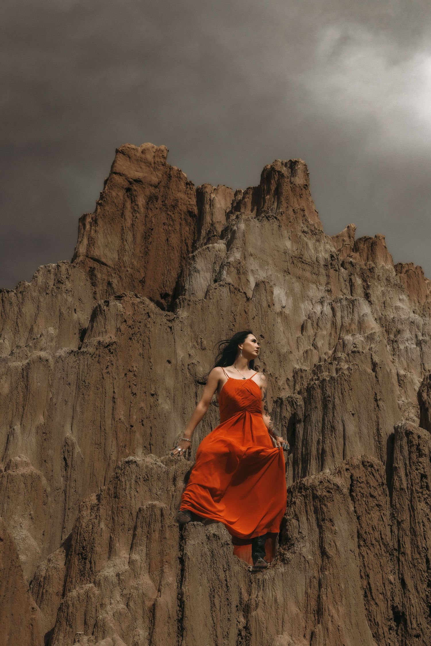Woman in flowing orange dress standing on rugged rocky cliffs under dramatic stormy sky.
