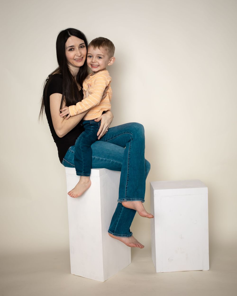Studio portrait showing a mother and child sitting on white pedestals against a neutral background.