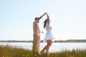 A couple in casual attire dances together on a scenic beach at sunset with water and reeds in the background.