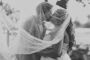 A romantic wedding couple embraces while a flowing veil billows in the breeze during an outdoor ceremony.