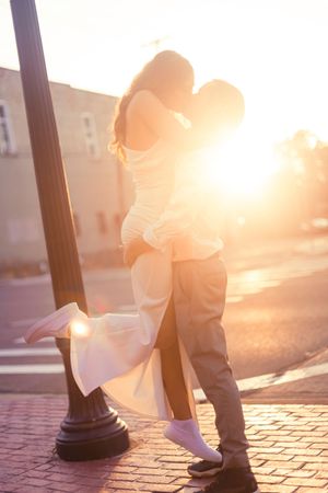 Two people's feet are silhouetted against a warm sunset on a cobblestone street with lamp posts.