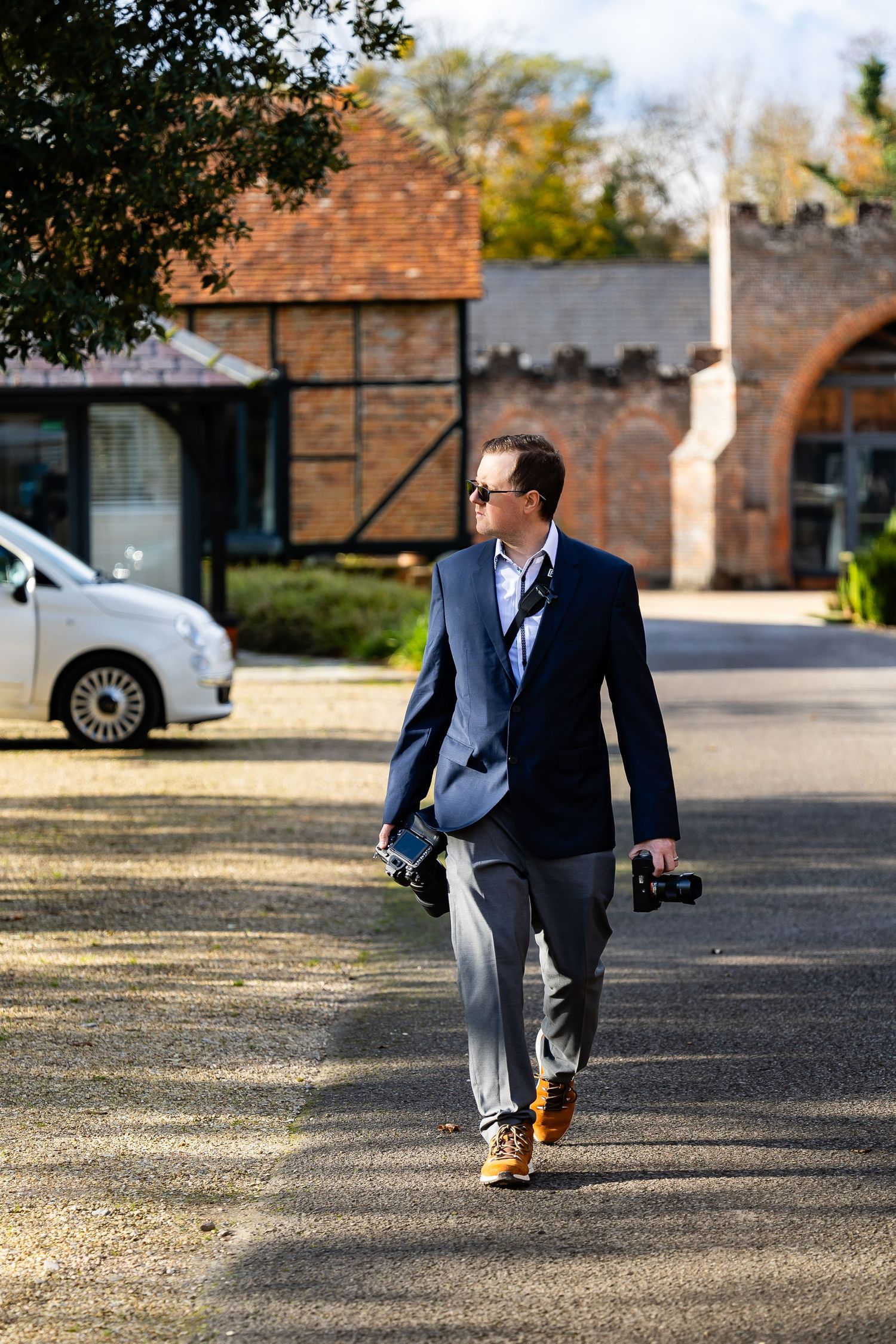 A person in a navy blazer walks through a courtyard carrying photography equipment.