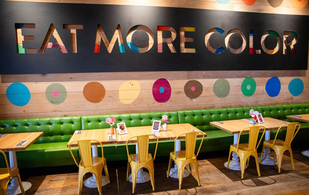 Modern restaurant interior with 'Eat More Color' text on wall, colorful dots, green banquettes and metal chairs.
