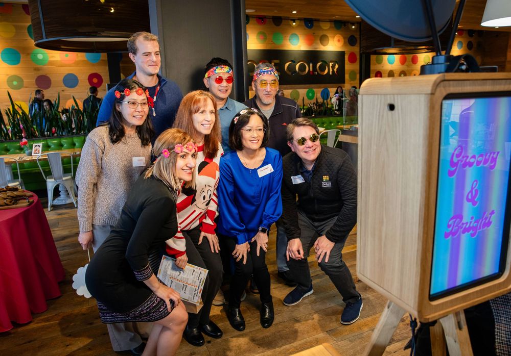 A group poses together at an indoor event with colorful decor and a digital display in the background.
