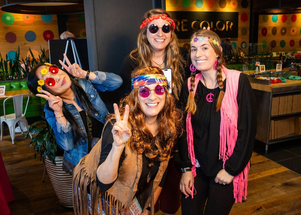 Party guests wearing colorful sunglasses and fringe fashion accessories pose together at a social gathering indoors.