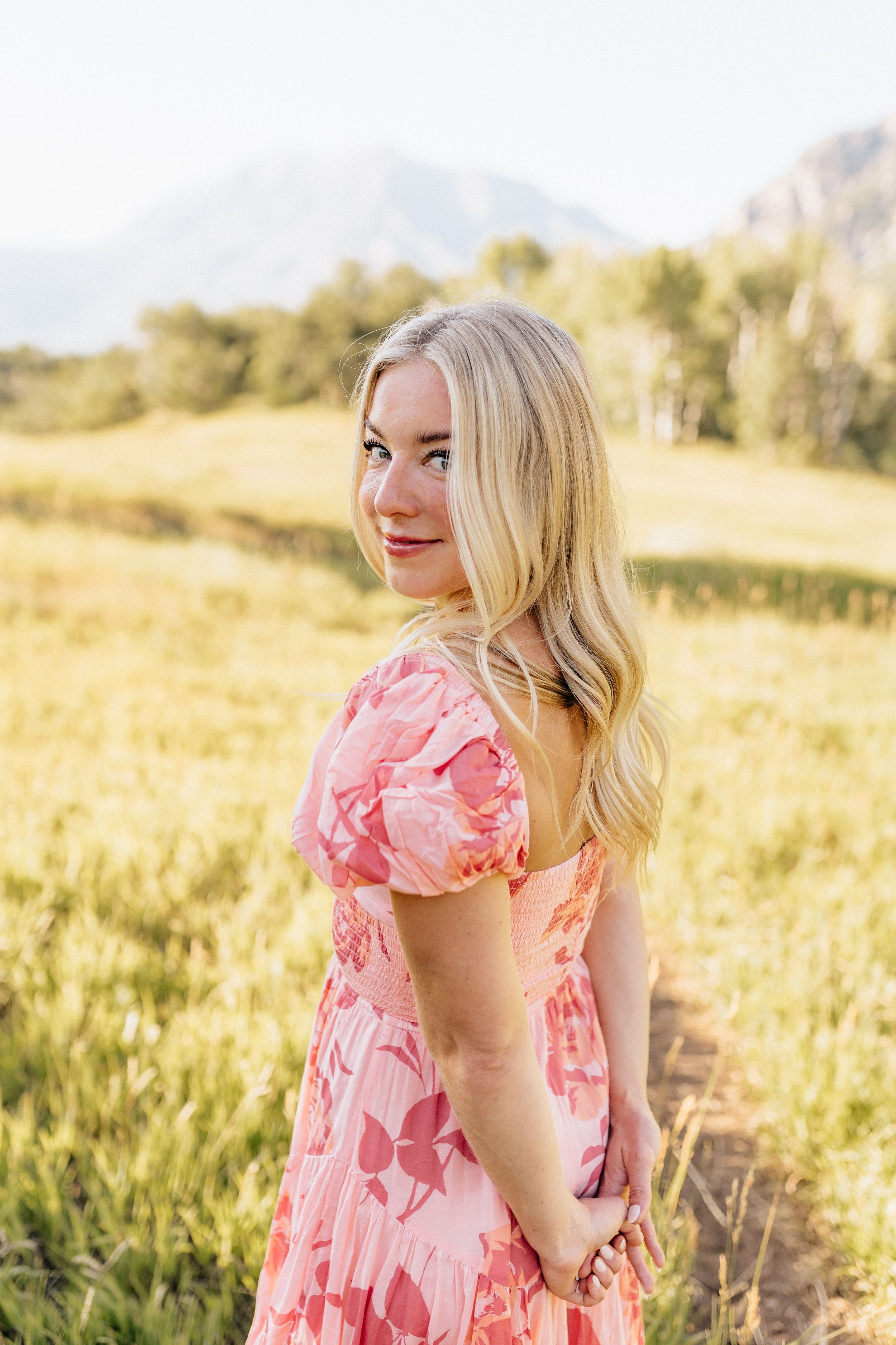 A woman in a pink floral dress poses in a golden mountain meadow at sunset.