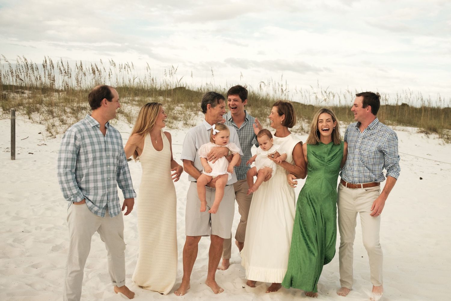 Family in light colored clothing shares tender moments on beach with seagulls flying overhead.
