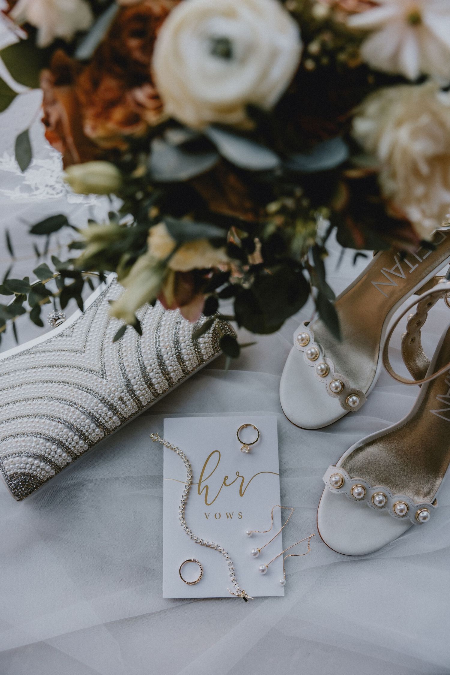Wedding details flatlay with white bridal shoes, rings, vows card, knit clutch and floral arrangement on tulle fabric.