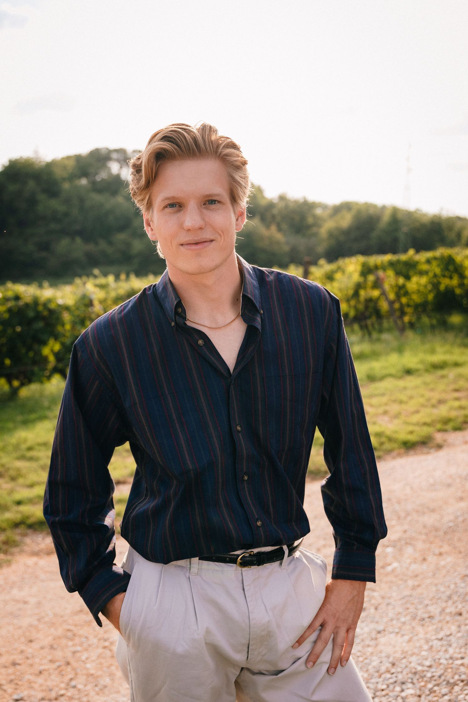 A person in a navy striped shirt poses casually in front of an Italian vineyard rows at sunset in Tuscany, Italy.