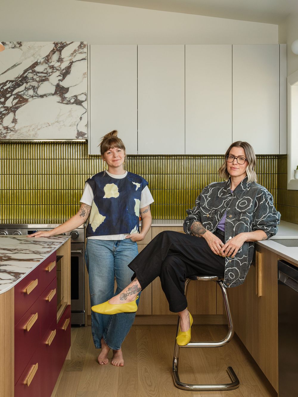 Two women pose in a modern kitchen with marble countertops, green tile backsplash, and burgundy cabinets.