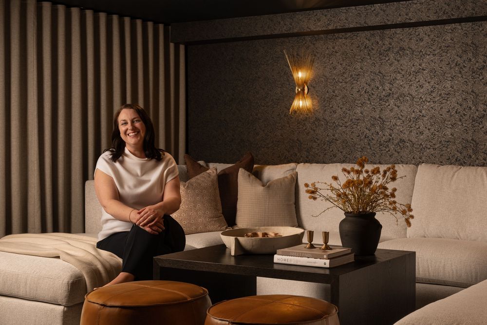 Woman smiling on a beige sectional sofa in a moody, elegantly designed living room with warm lighting.