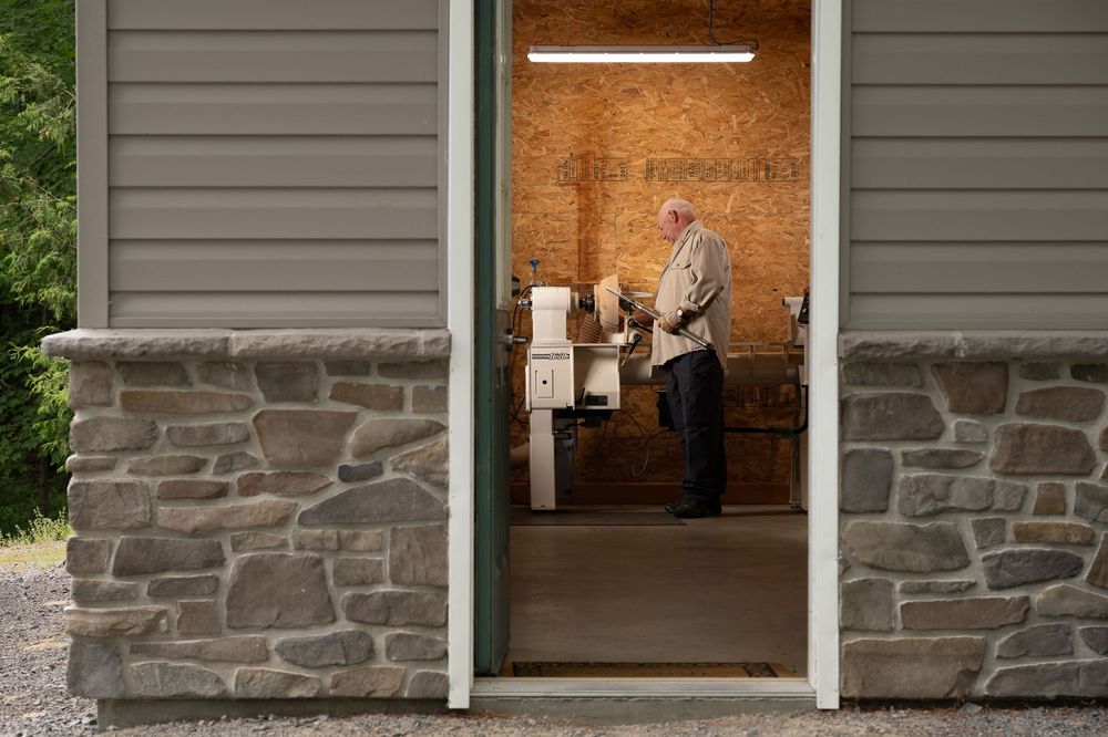 A wood turner creates a piece in a doorway with stone foundation and gray siding exterior.
