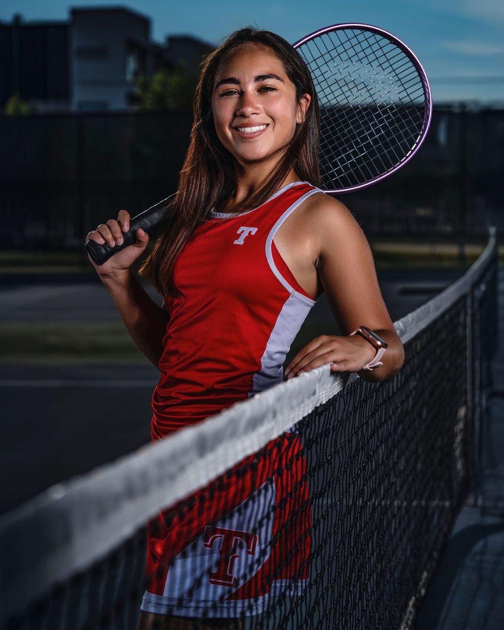 Tennis player in red uniform poses with racquet against chain link fence during evening photoshoot.