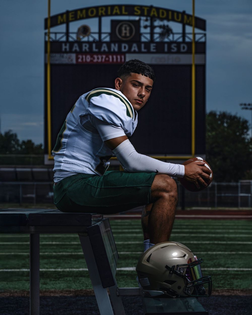 Football player sitting alone on sideline bench at Memorial Stadium during evening hours with sports field visible behind.