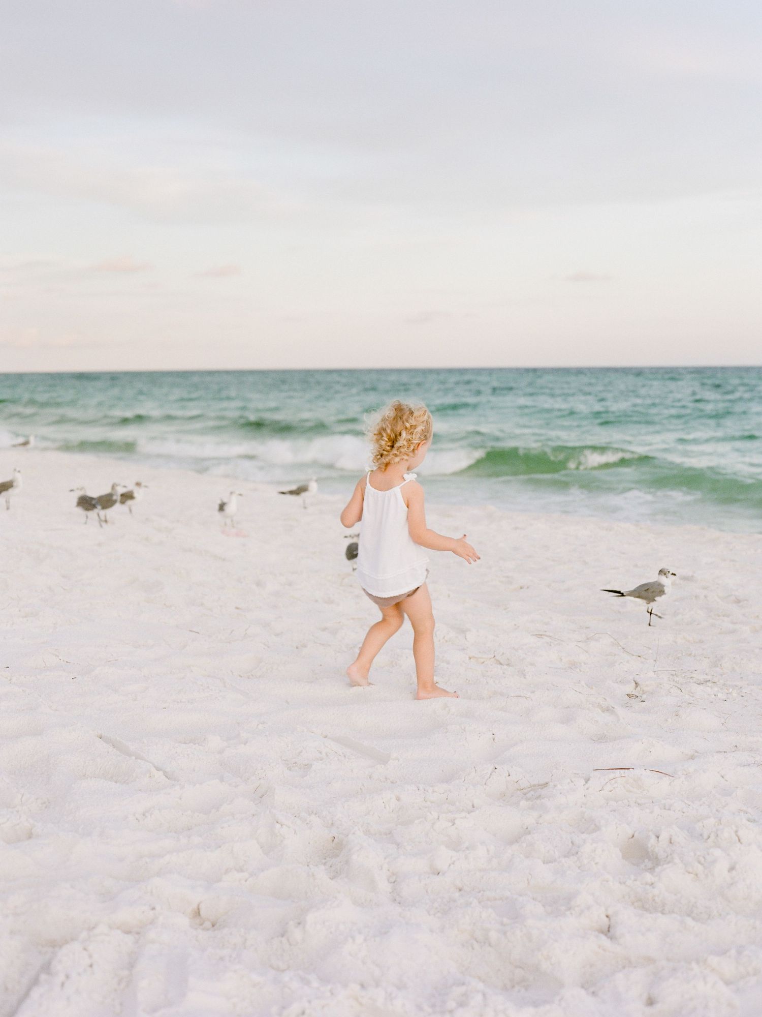 Young child in a white dress stretches their arms toward the sky in a joyful pose during sunset.