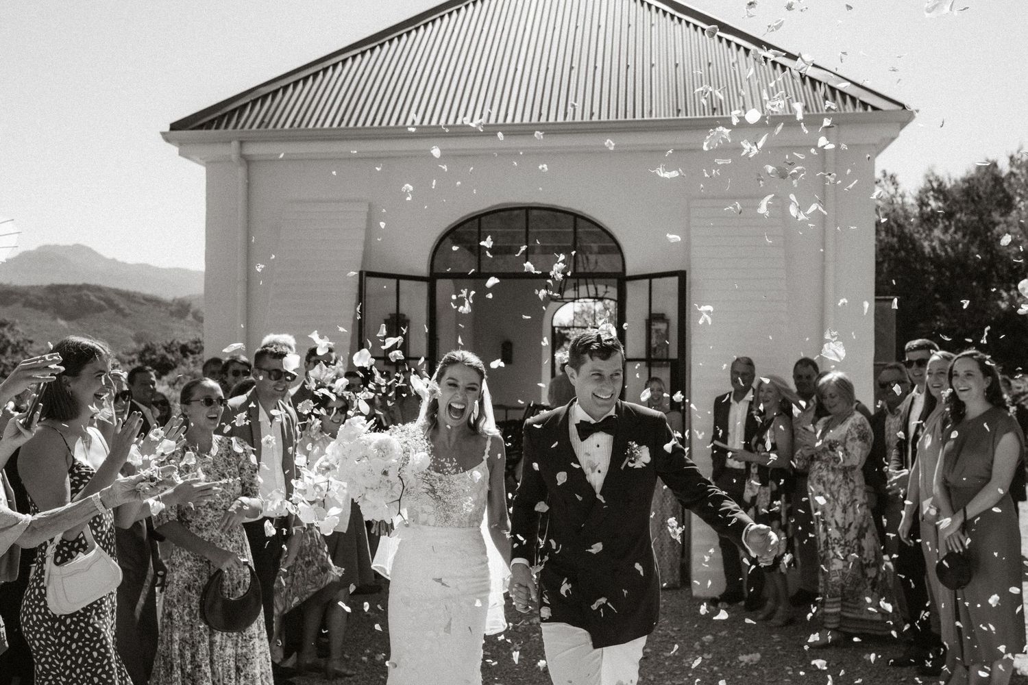 A black and white wedding photo with flower petals being thrown over the celebrating couple.