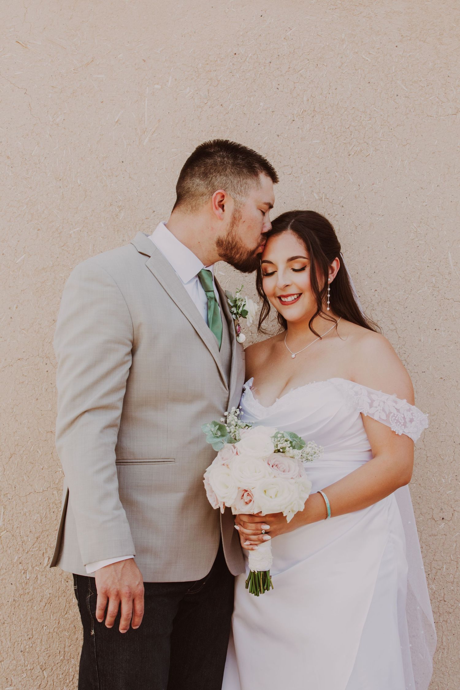 Bride and groom share a tender moment, groom kissing bride's forehead, bride holding white floral bouquet against beige wall.