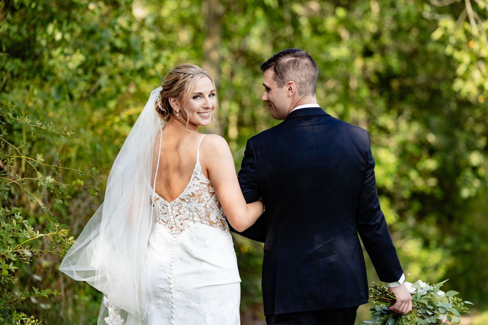 Bride in backless lace gown with veil smiles over shoulder as groom holds her hand outdoors.