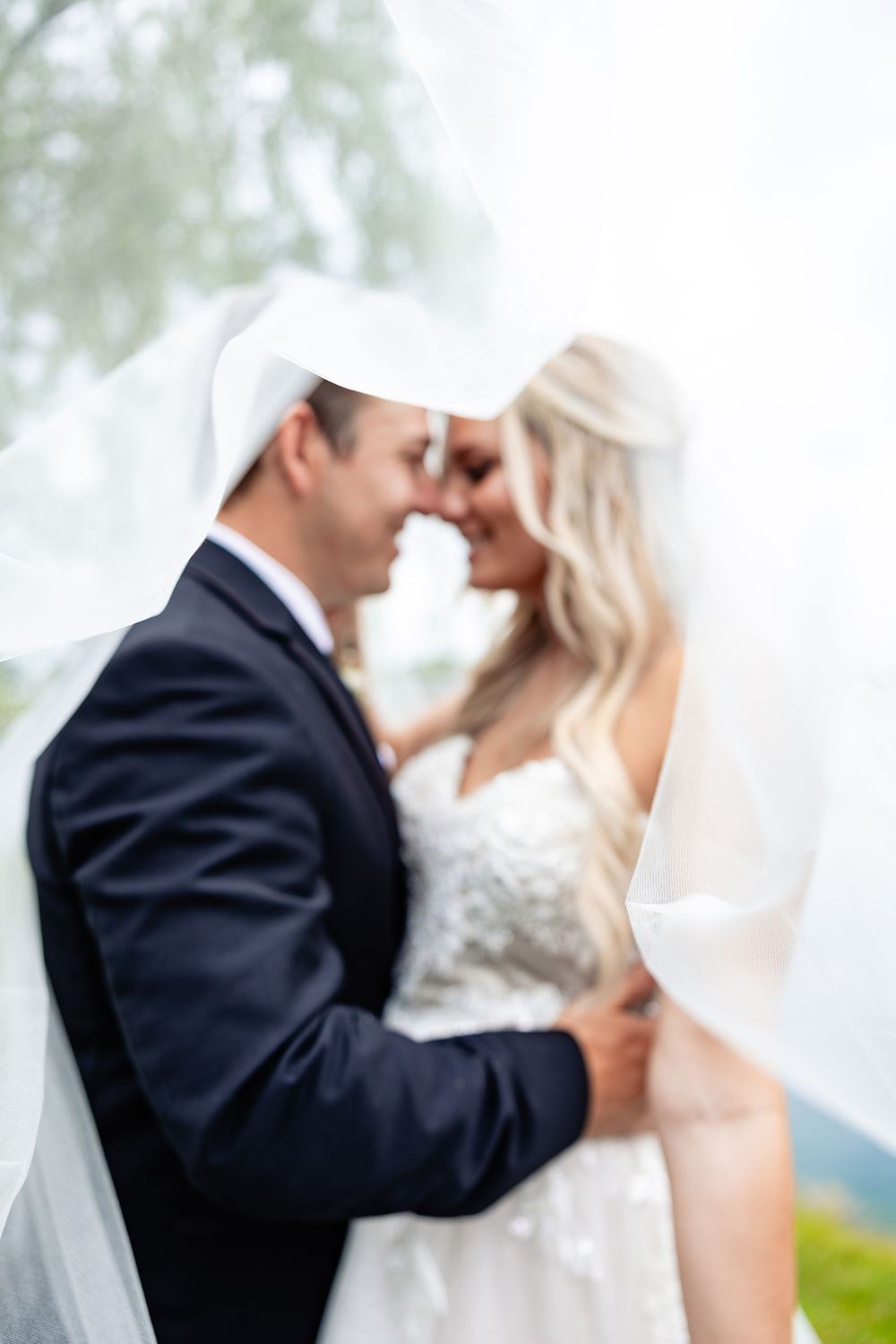 Romantic blurred photo of bride and groom touching foreheads under flowing white veil outdoors on wedding day.