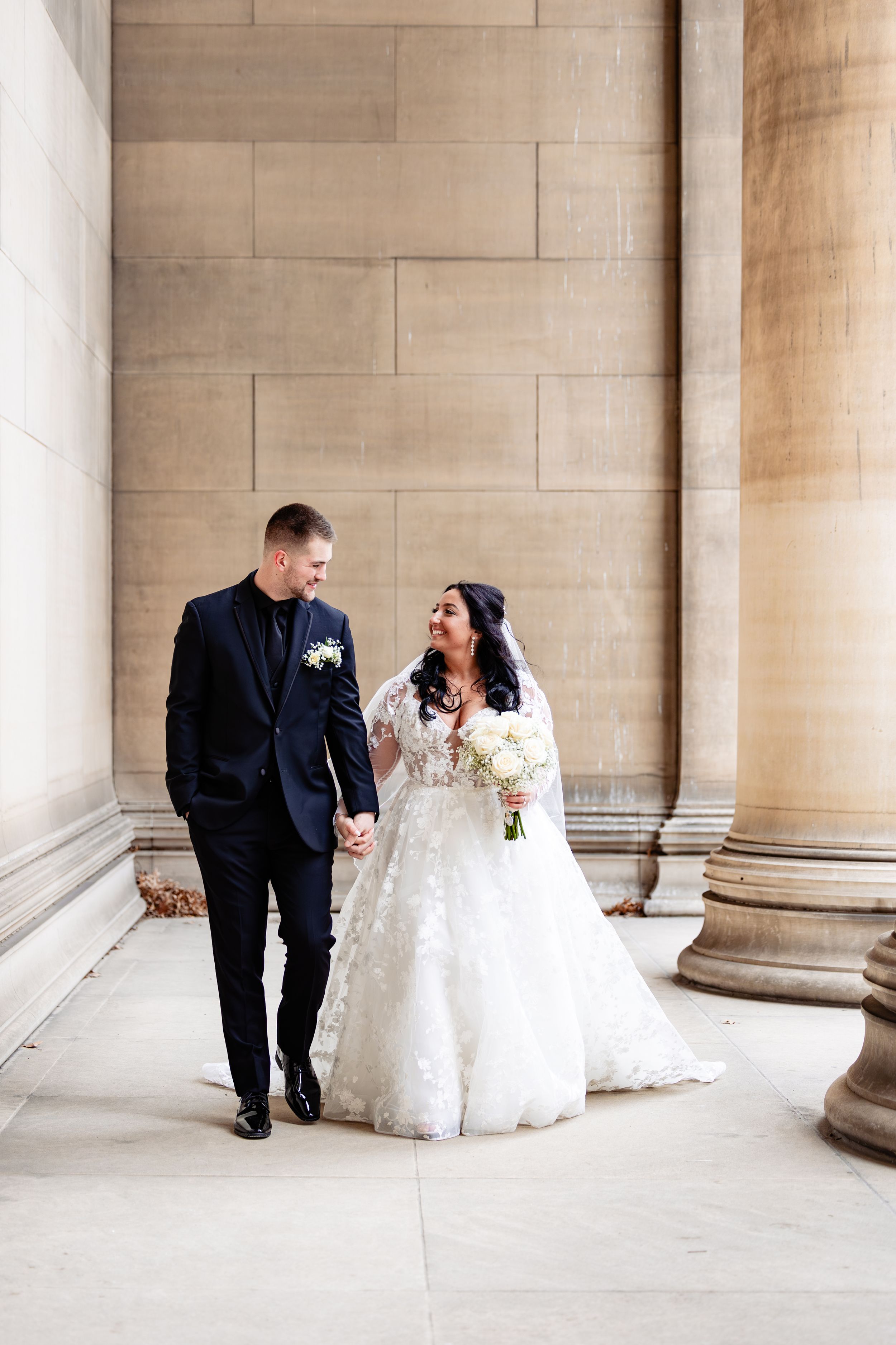 Bride and groom walk hand in hand past grand stone columns, sharing a joyful glance on their wedding day.
