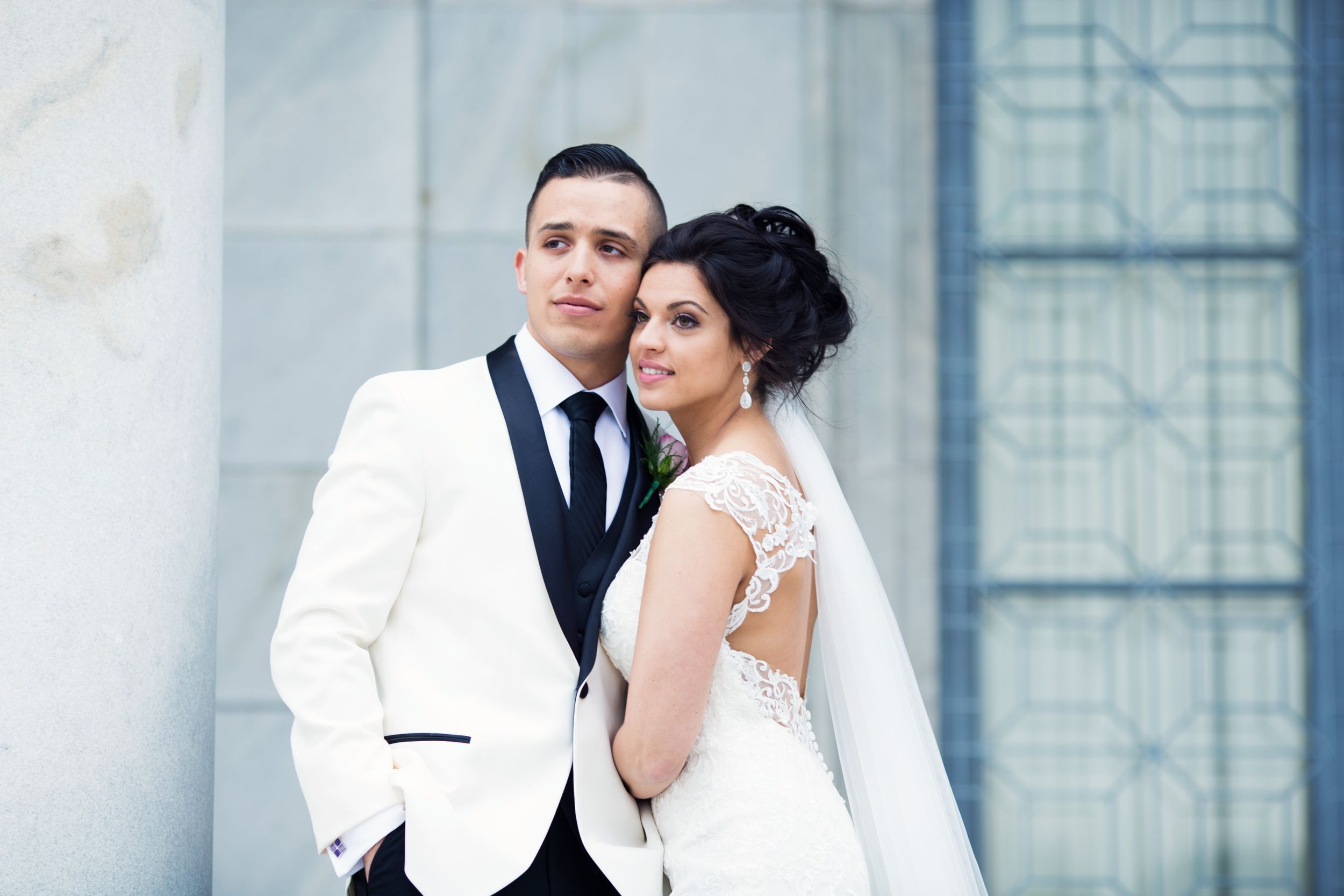 Newlyweds posing elegantly outdoors, groom in white tuxedo and bride in sleek white gown with veil.