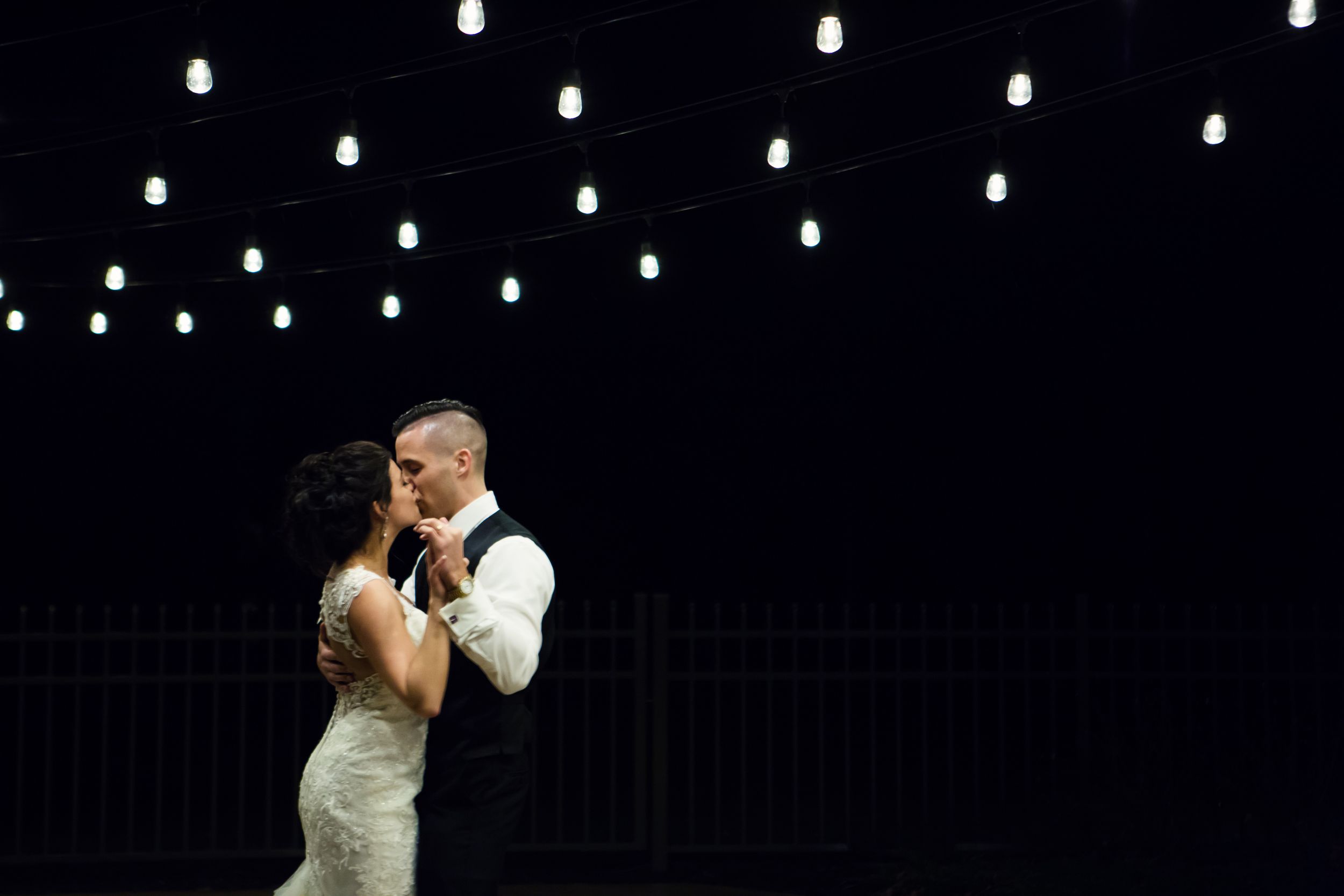 Bride and groom sharing first dance under warm string lights against a dramatic dark background.