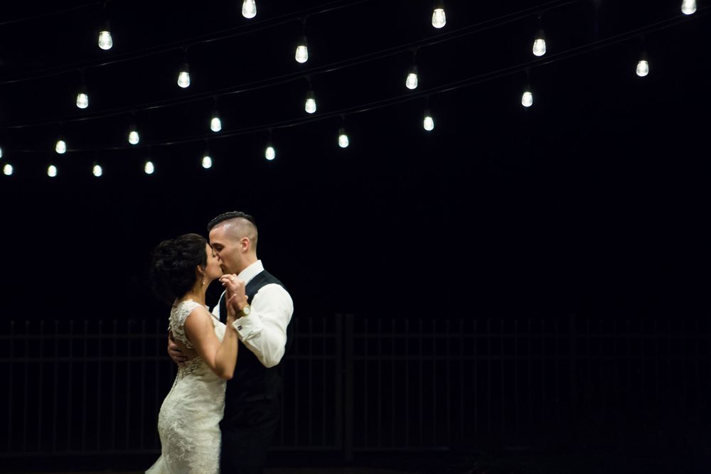 Bride and groom sharing first dance under warm string lights against a dramatic dark background.
