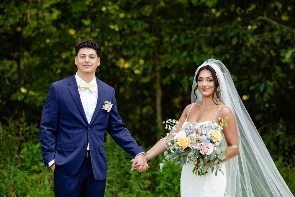 Bride in white gown and groom in navy suit hold hands outdoors among lush greenery.