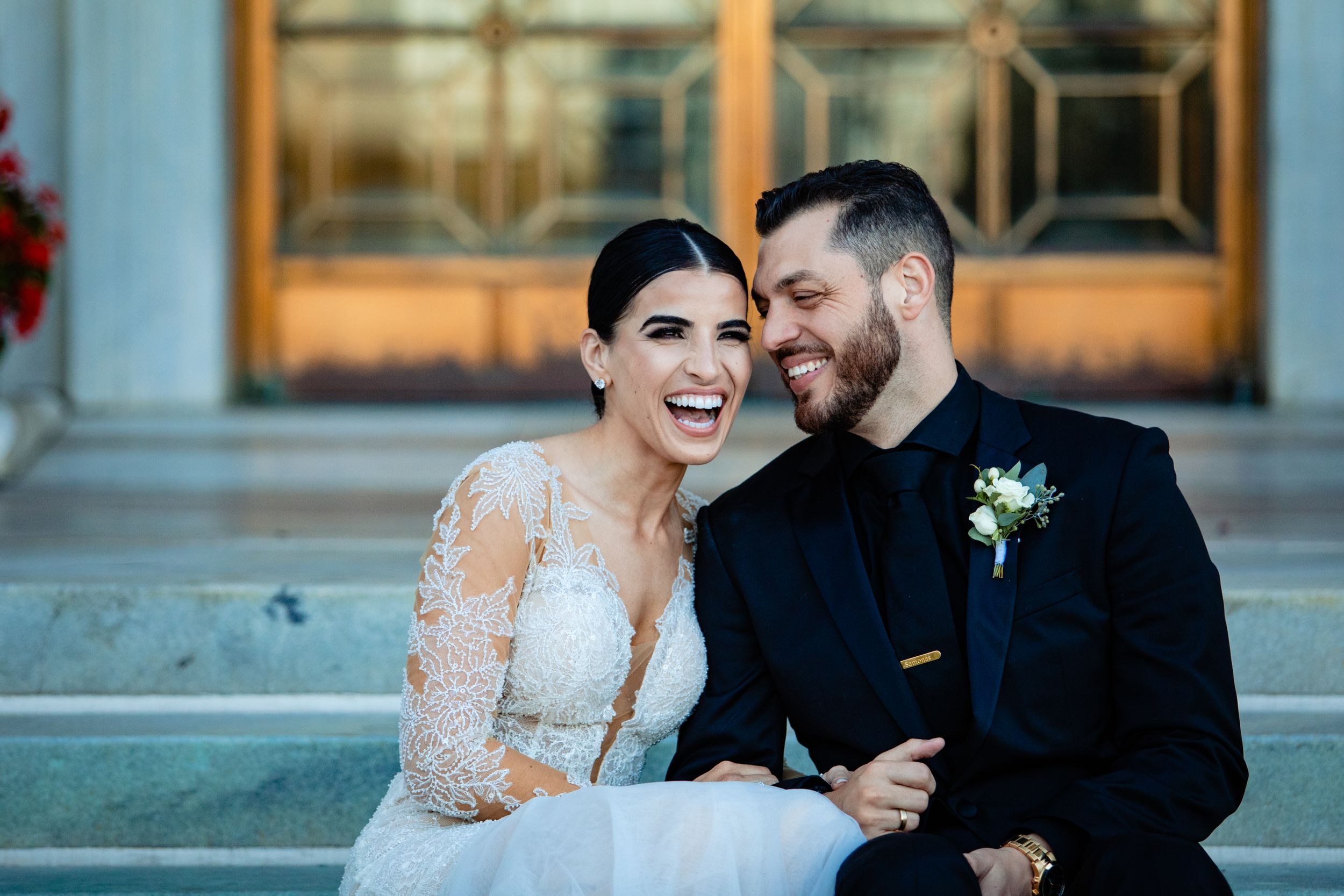 Laughing bride and groom sitting on steps, bride in lace long-sleeve gown, groom in black suit with boutonniere.