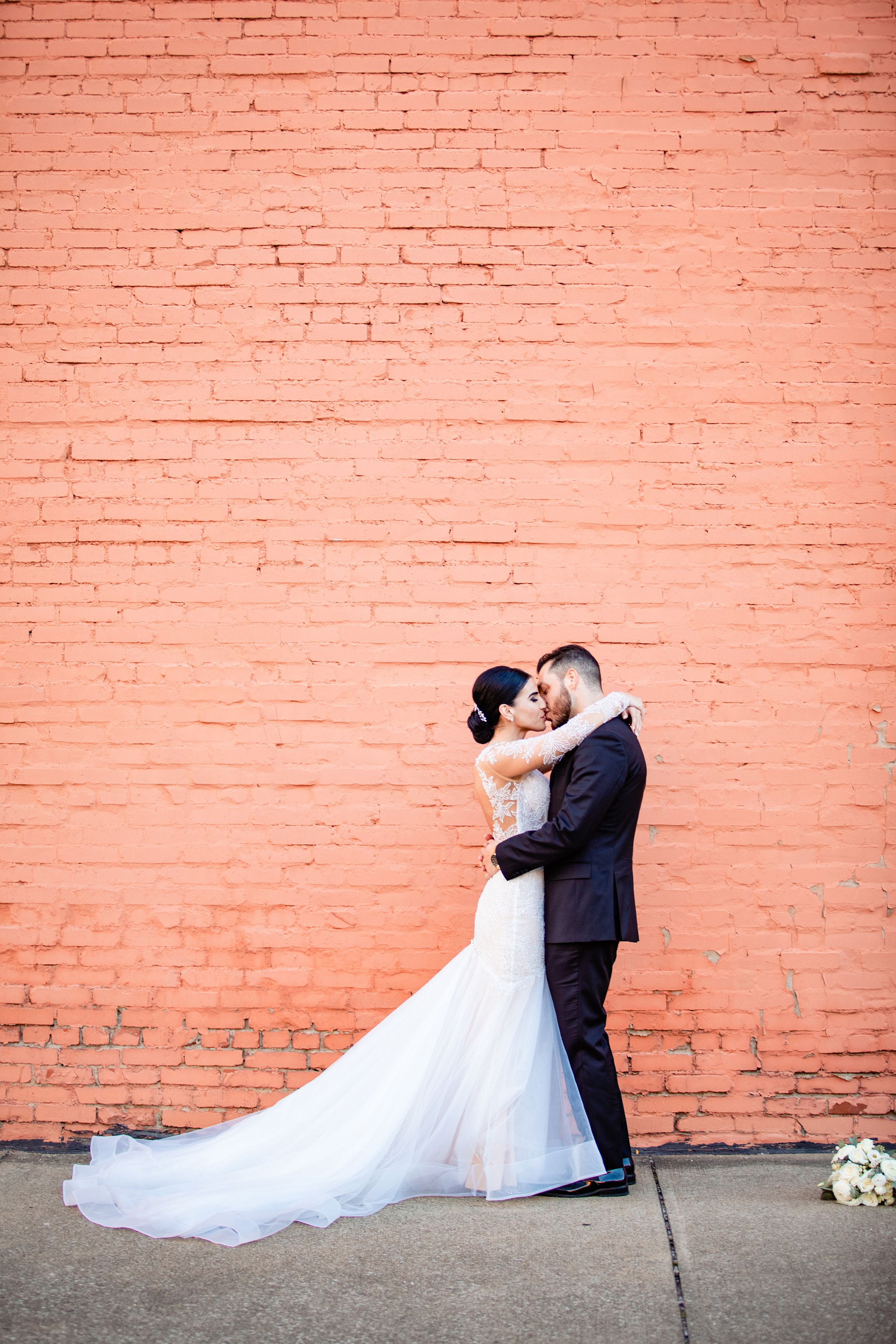 Bride and groom embracing against a coral brick wall, bride in flowing white gown with long train.