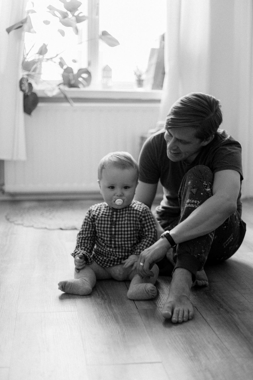 Mustavalkoinen dokumentaarinen perhekuva jossa isä ja vauva istuvat lattailla kotona ikkunan edessä vauvan katsoessa kameraan.