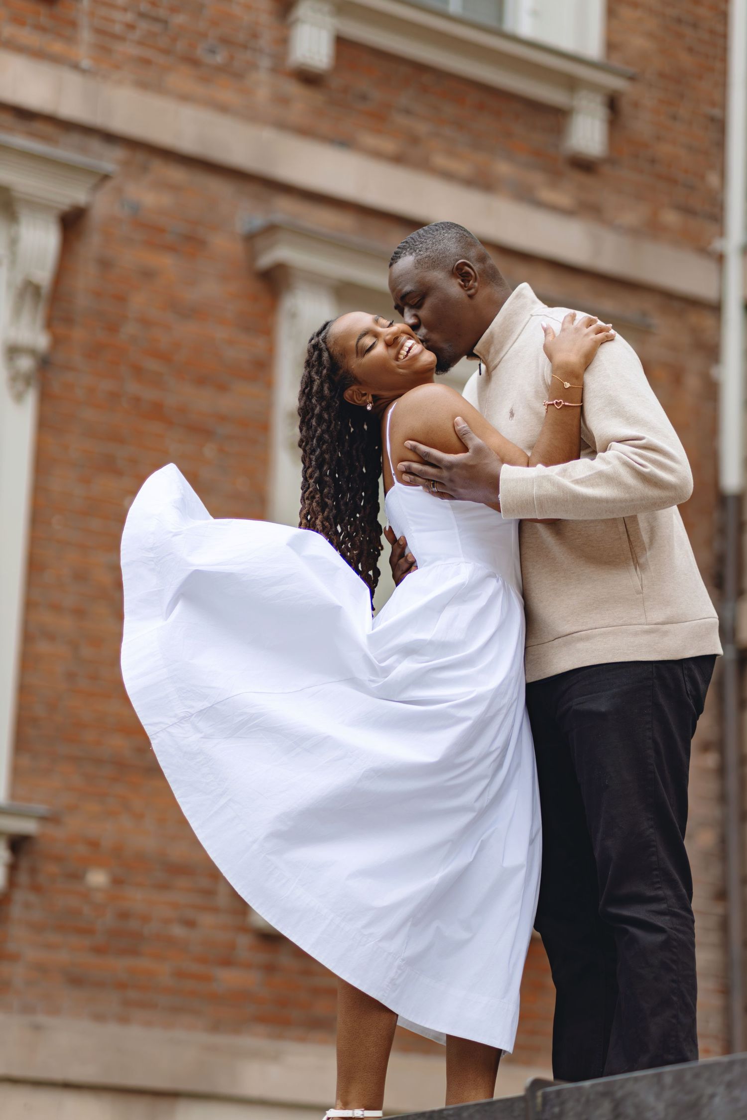Groom kisses bride in flowing white dress outside brick building in romantic urban setting.
