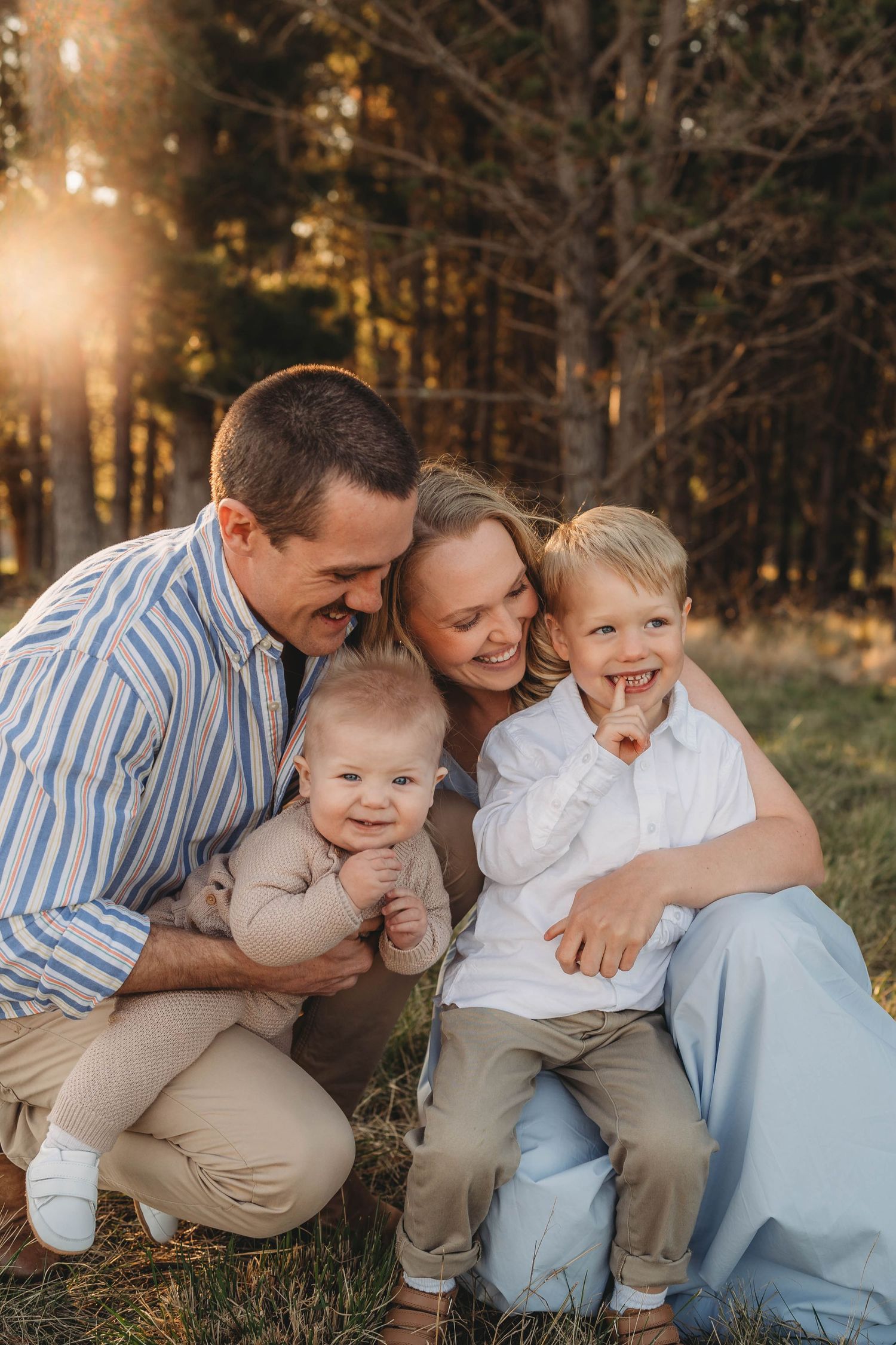 Happy family of four posing outdoors at golden hour, parents smiling with baby and toddler son in a sunlit forest.