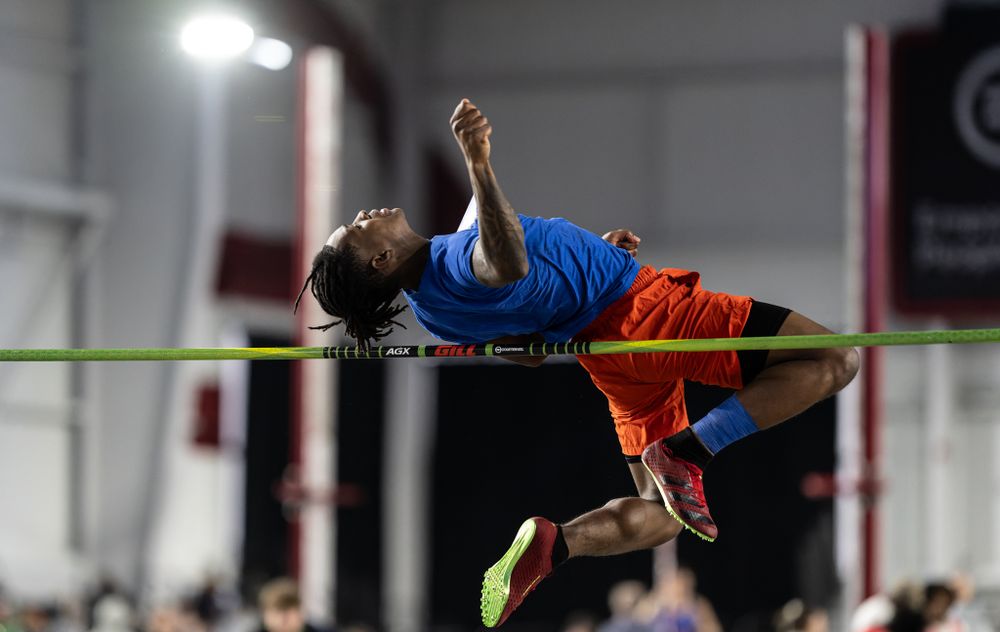 High jumper in blue jersey and orange shorts arches backwards over high jump bar during indoor competition.