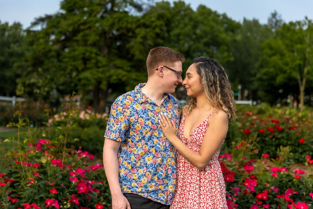 Two figures stand together in a vibrant rose garden wearing colorful floral prints on a sunny day.