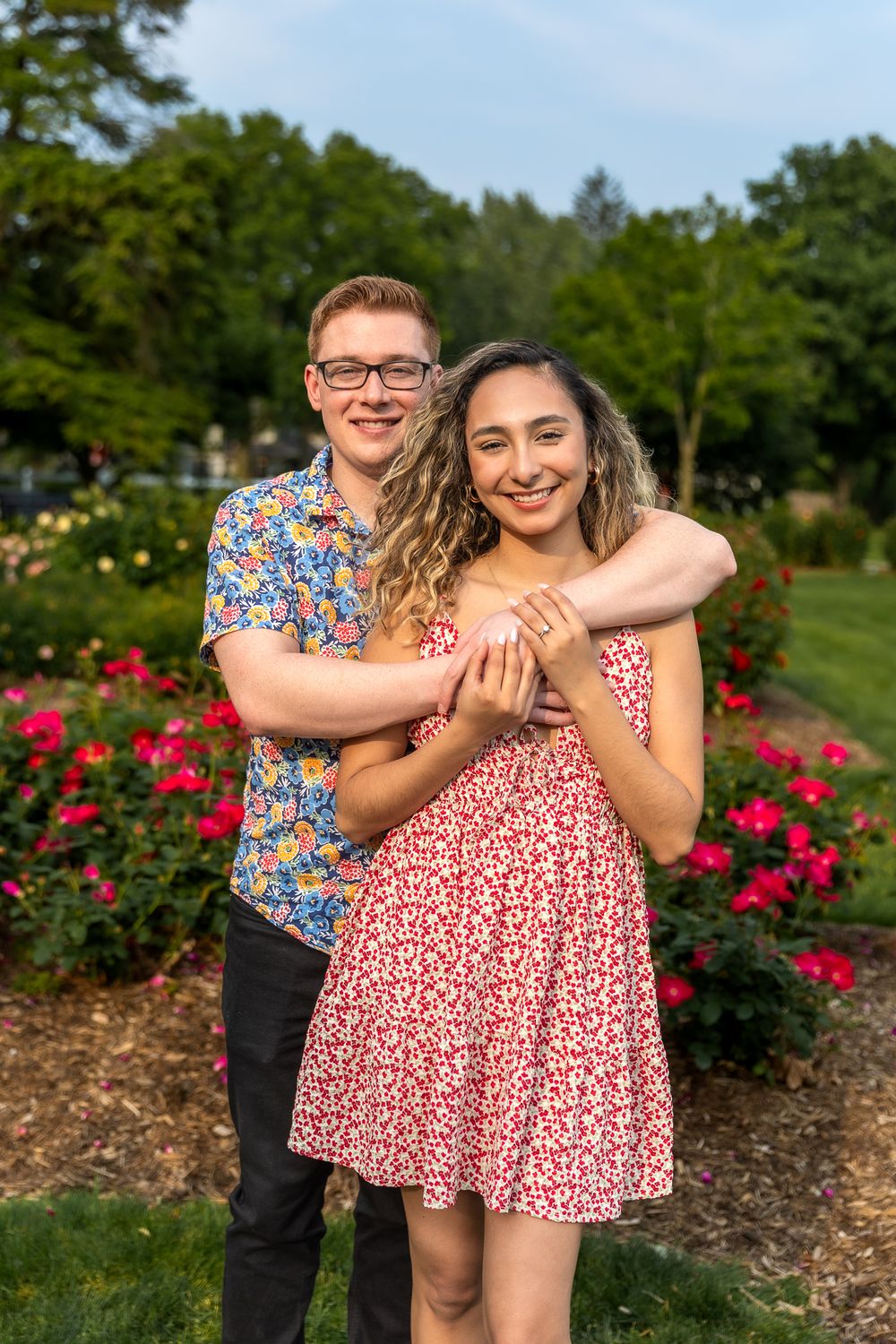 A joyful couple pose together in a garden with blooming red roses on a sunny day.