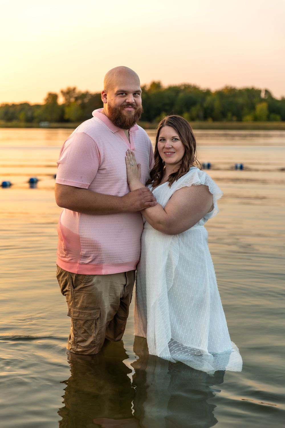 A couple embraces in shallow water during sunset, wearing pink shirt and white dress, creating romantic silhouettes.