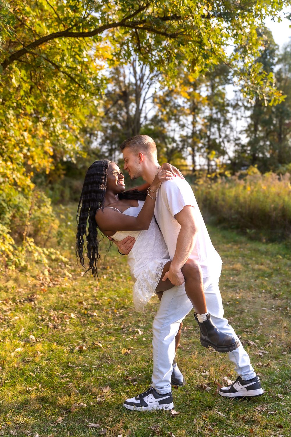 A playful couple in white clothing share a romantic dip pose against fall foliage.