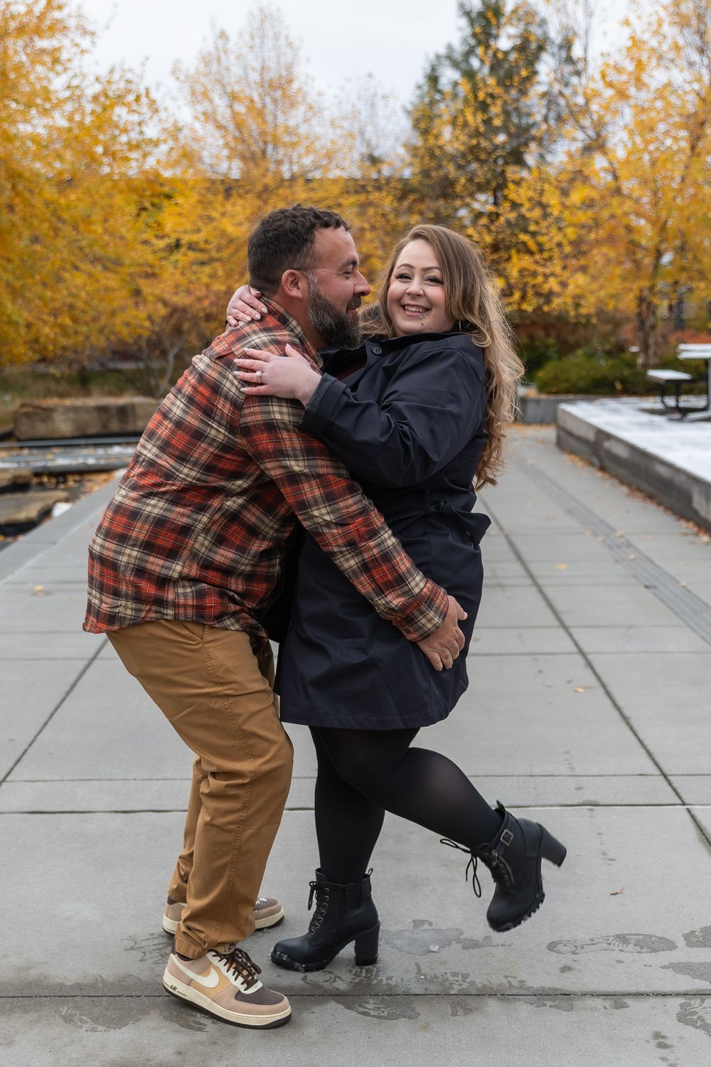 A couple shares a playful moment dancing together on a concrete path surrounded by autumn trees with golden leaves.