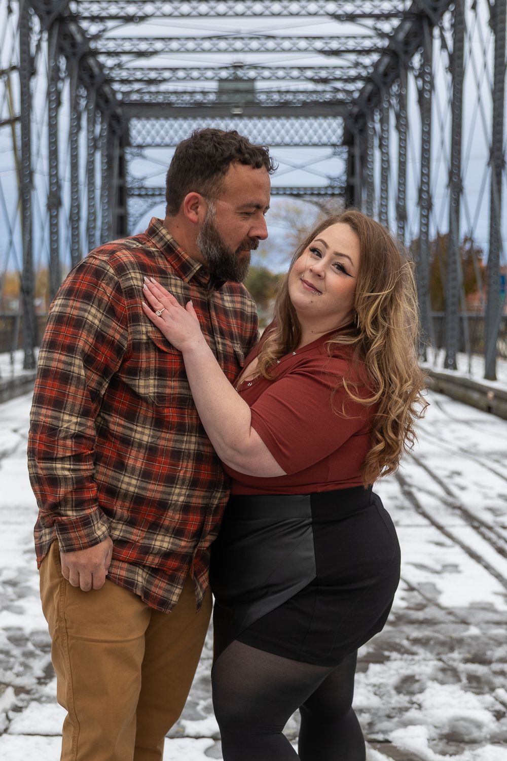 A romantic couple embraces on a snowy railroad bridge, with the woman wearing red and the man in a plaid flannel shirt.