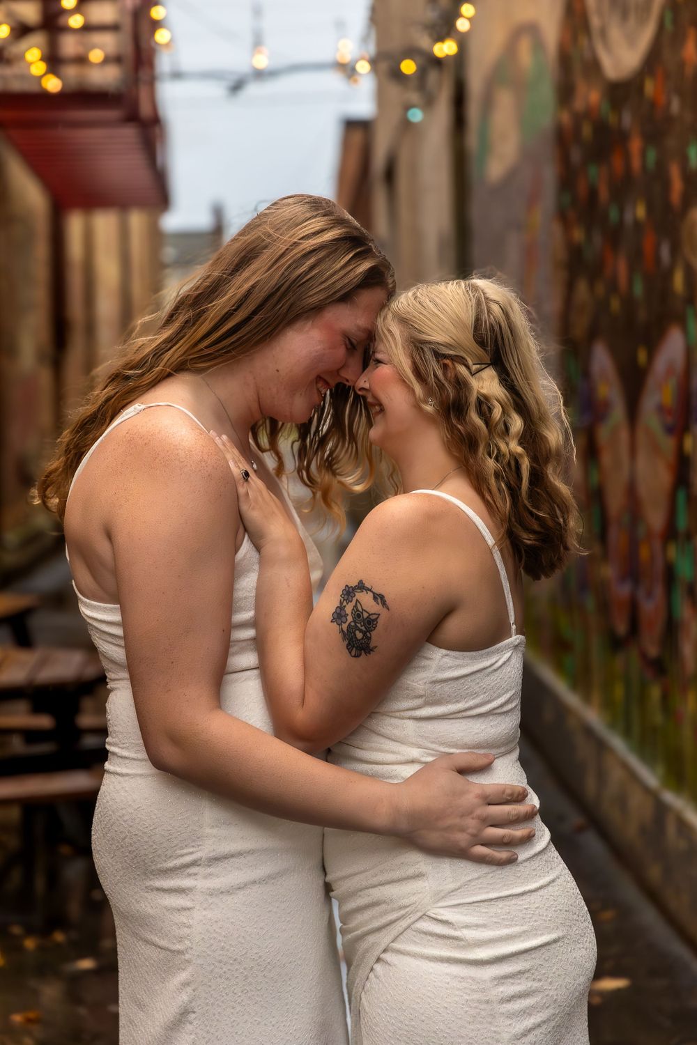 Two people in white dresses share an intimate moment in an alley decorated with string lights.