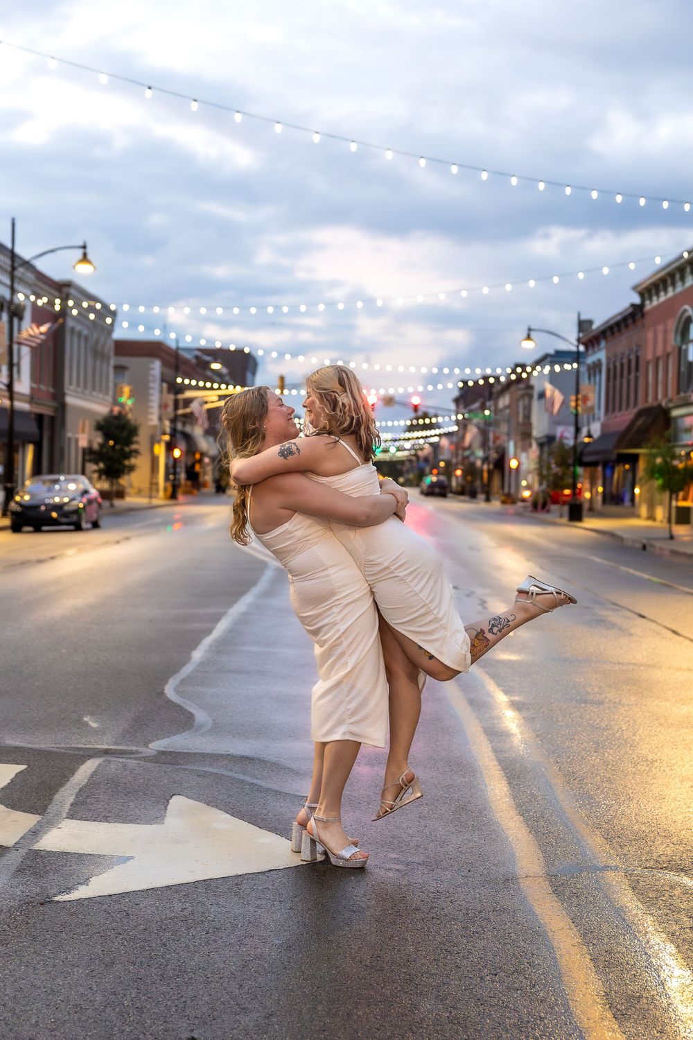 Happy couple in white dresses embracing on city street at dusk with twinkling string lights overhead.