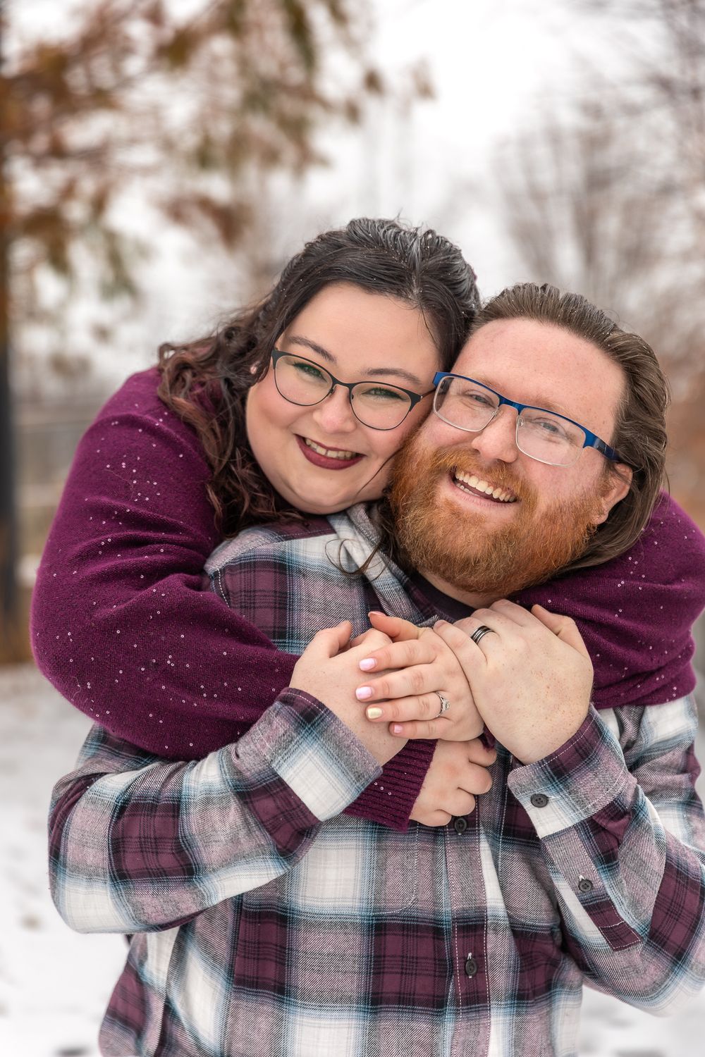 A couple wearing plaid and purple embraces while laughing in the snow during a winter photo session.