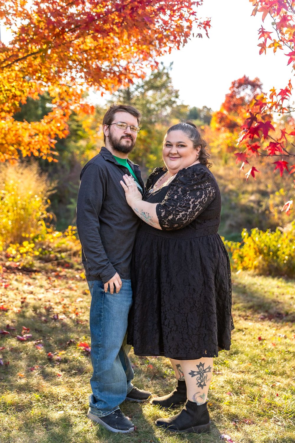 A couple poses in front of vibrant autumn foliage wearing black and casual attire.