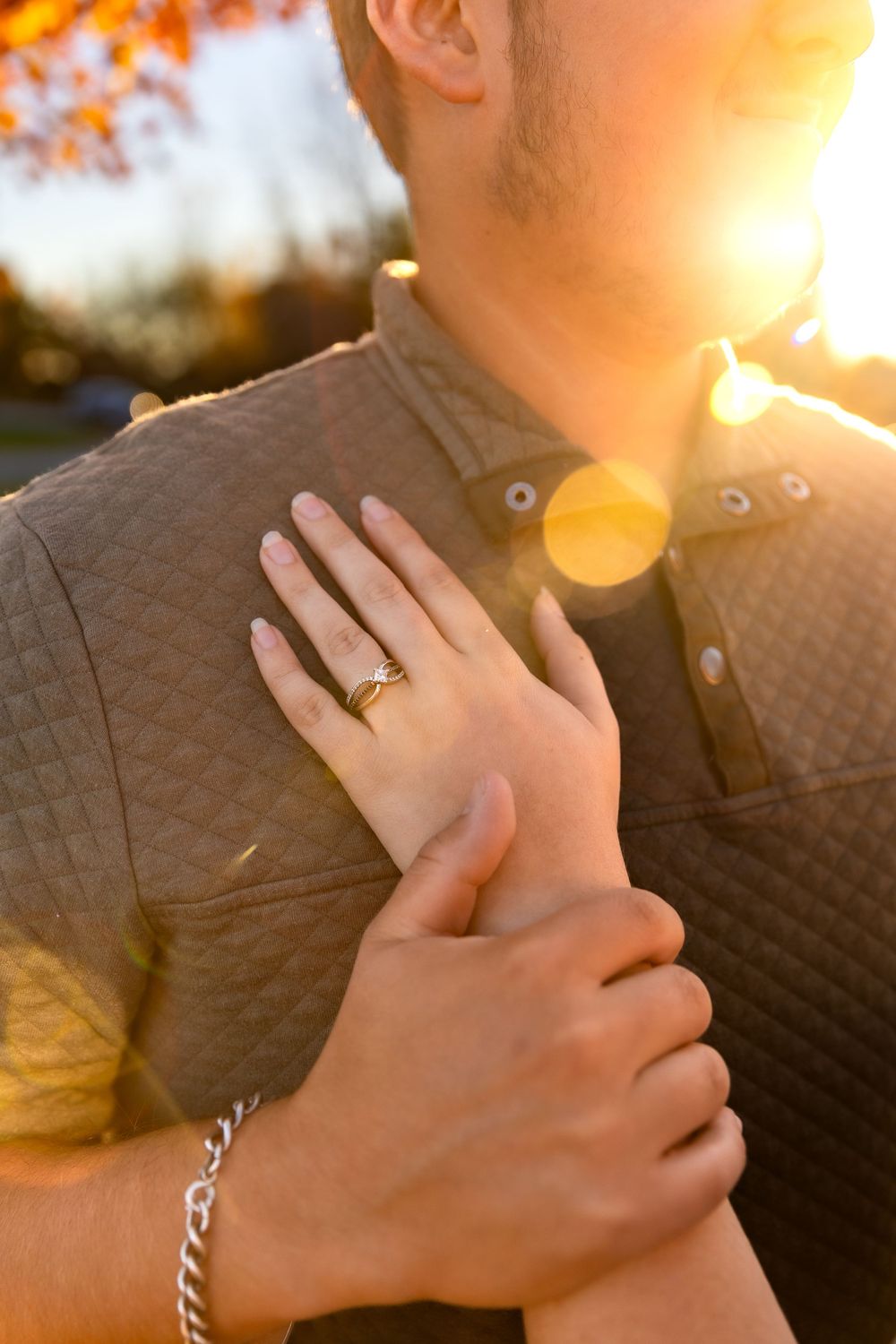 Hands showing off an engagement ring with sun flare and autumn colors in background.