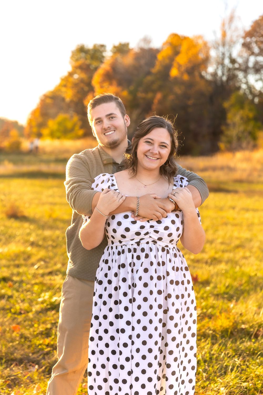 A couple embraces in a field during golden hour with fall foliage behind them.